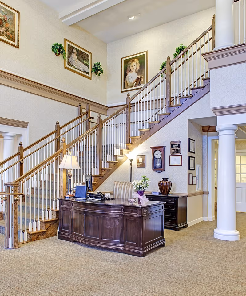 Interior view of a senior living facility lobby with a wooden reception desk, a striped chair, a table lamp, and a vase with flowers. Behind the desk is a staircase with wooden handrails and white balusters leading to an upper floor. The walls are decorated with framed paintings and greenery. There are columns and a carpeted floor.