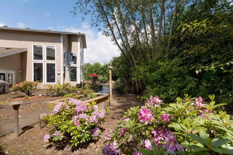 Outdoor garden area with blooming pink flowers and green shrubs near a beige building with large windows. A paved walkway leads through the garden, and there is a red umbrella visible in the background under a partly cloudy sky.