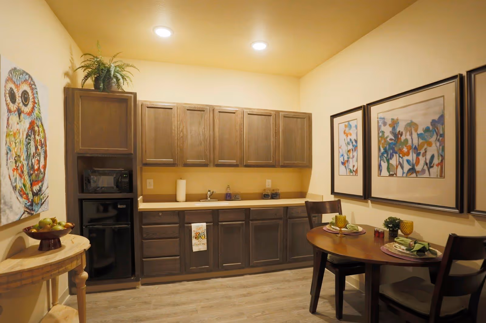 A small kitchen and dining area with wooden cabinets, a countertop with a sink, a microwave, and a mini refrigerator. There is a round wooden dining table set for two with plates, glasses, and napkins. The walls are decorated with colorful framed artwork and a painting of an owl. A small wooden side table holds a bowl of fruit and a potted plant.