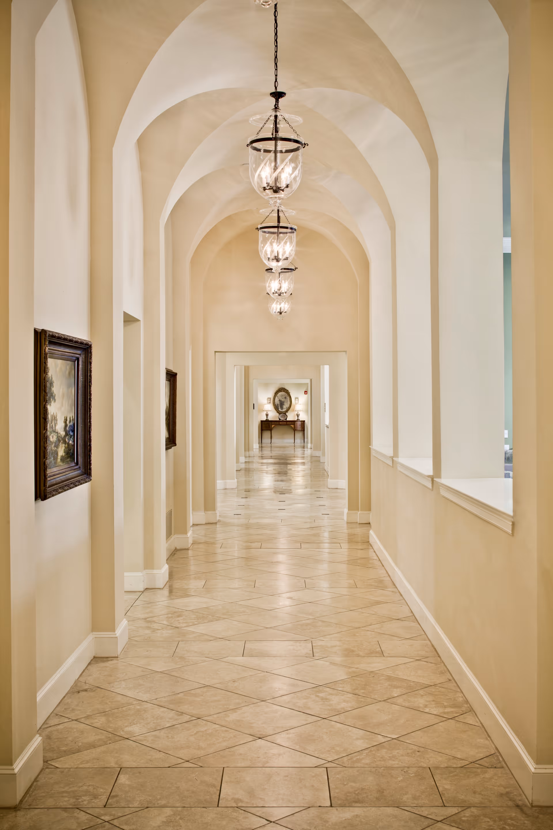 A long, elegant hallway with arched ceilings and hanging lantern-style light fixtures. The walls are painted a soft beige color, and there are framed paintings on the left wall. The floor is tiled with large, light-colored tiles arranged in a diagonal pattern. At the end of the hallway, there is a table with decorative items and a framed portrait above it.