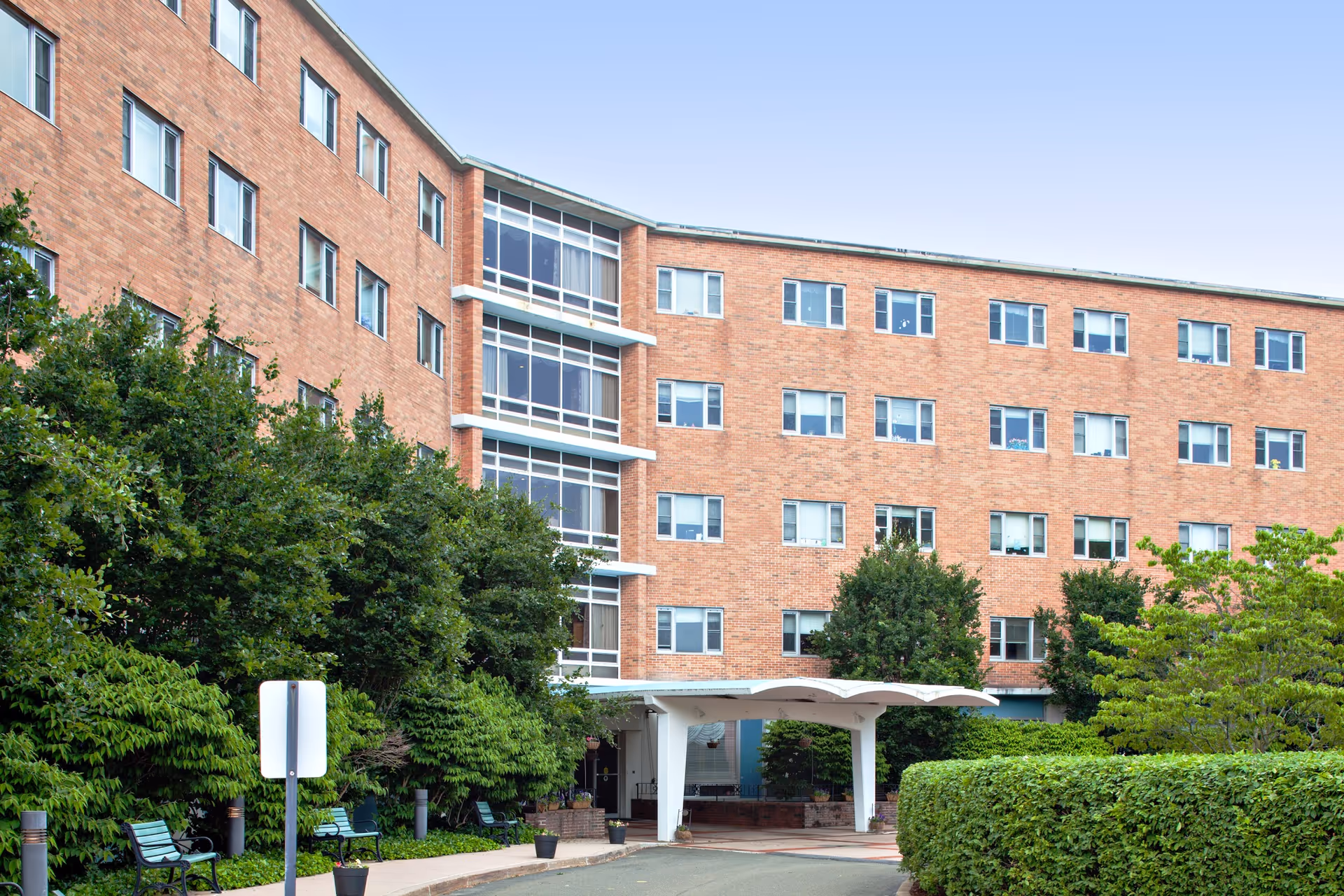 Exterior view of a multi-story brick building with many windows, surrounded by green trees and bushes. There is a covered entrance with a white canopy and benches along the sidewalk leading to the entrance.