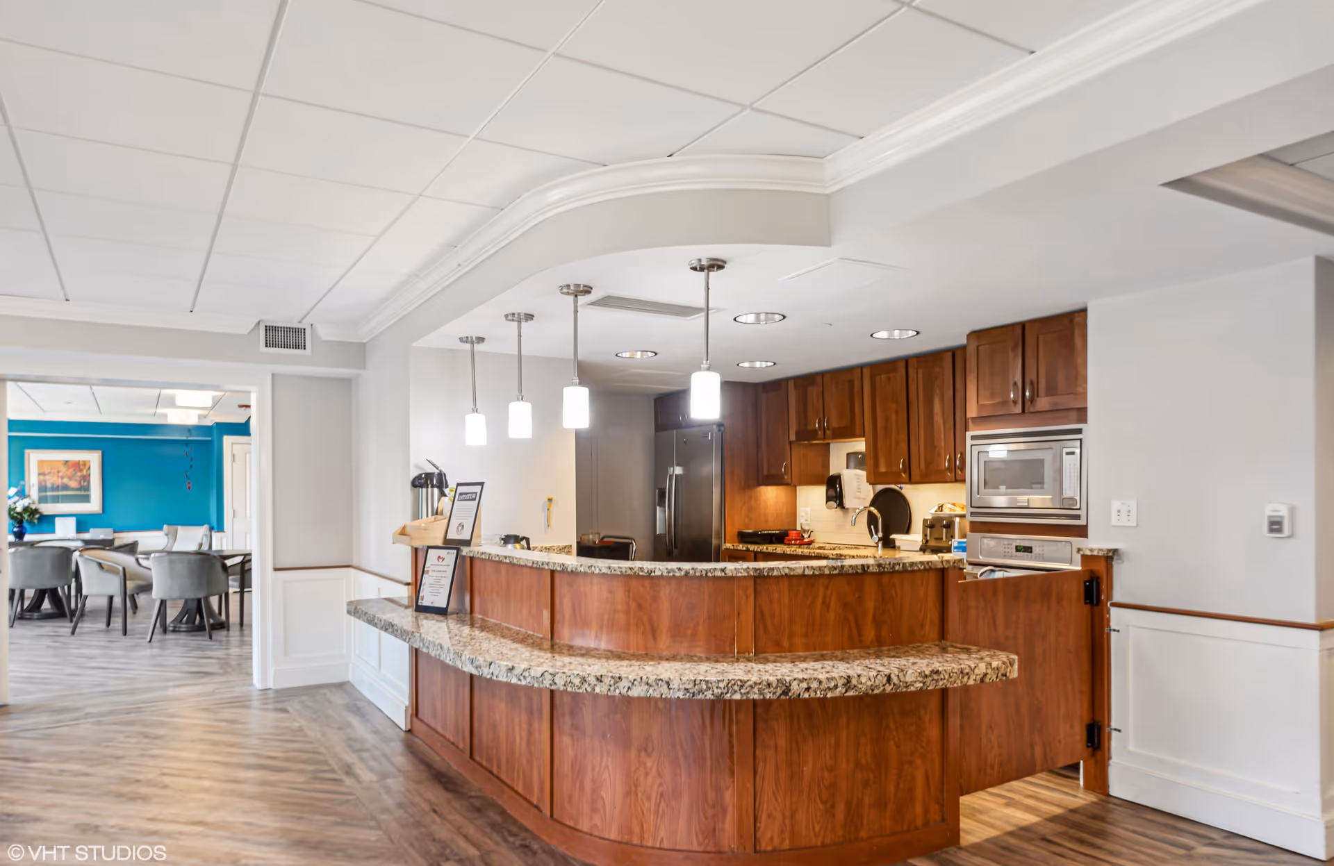 Interior view of a senior living facility kitchen area with wooden cabinets, granite countertops, and modern pendant lights hanging from the ceiling. In the background, there is a dining area with tables and chairs, and a bright blue accent wall with framed artwork.