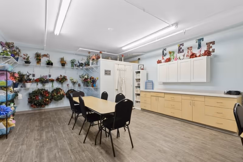 Craft room with a central table and chairs, storage cabinets, and shelves filled with floral arrangements and wreaths.