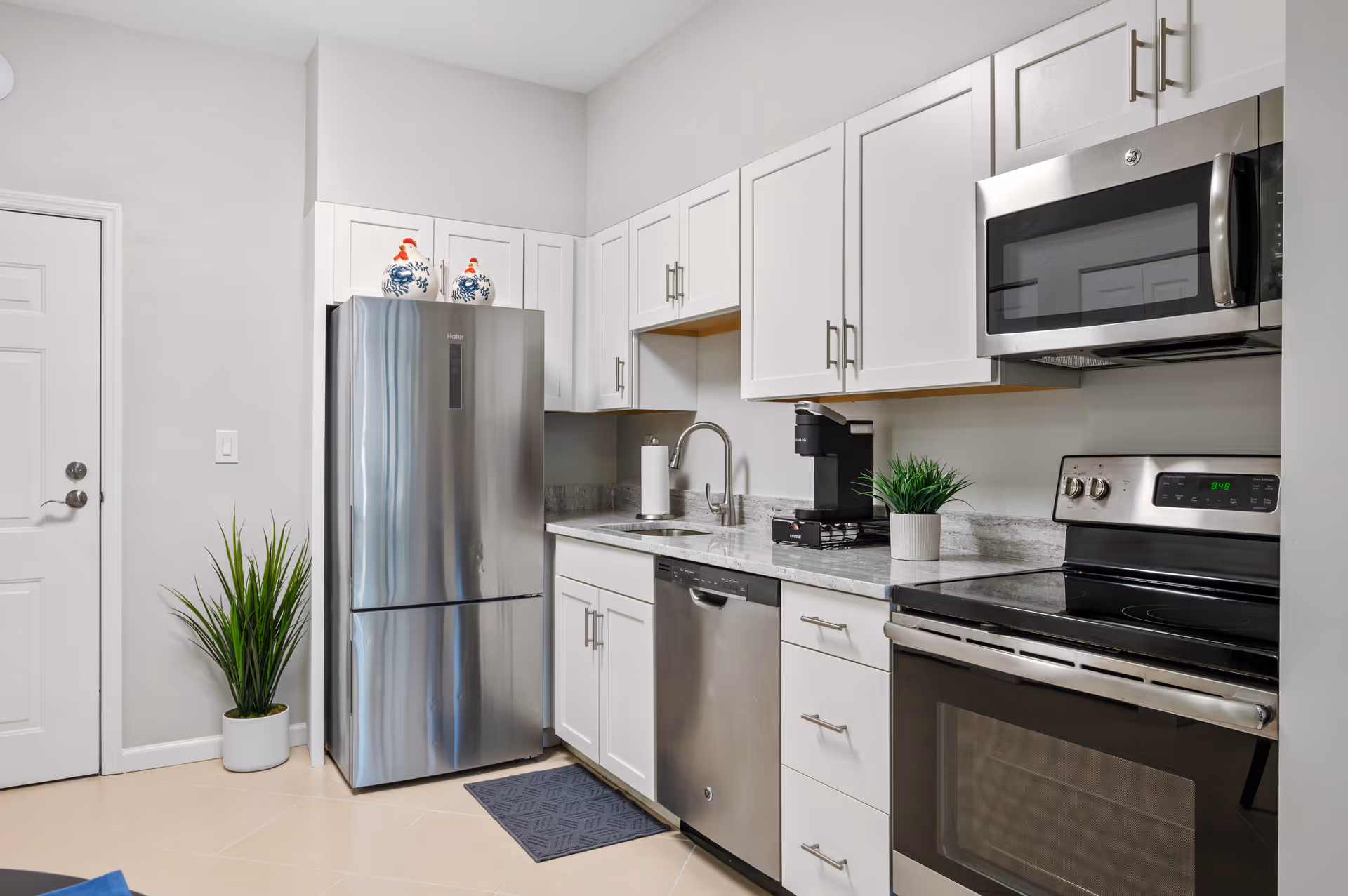 Modern kitchen with white cabinets, stainless steel refrigerator, dishwasher, microwave, and electric stove. There is a small green plant on the countertop, a coffee maker, and two decorative ceramic roosters on top of the refrigerator. A potted plant is placed on the floor near a white door.