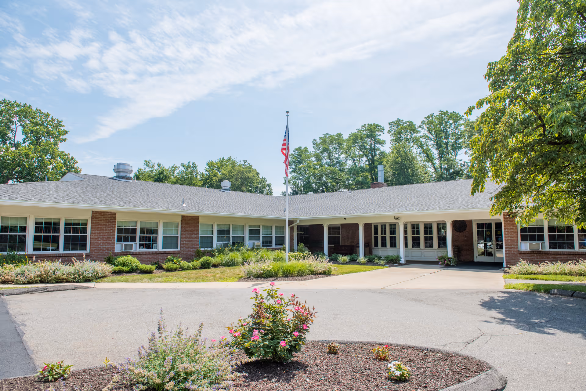Exterior view of a single-story brick building with multiple windows, a central entrance, and an American flag on a flagpole in front. The building is surrounded by greenery, bushes, and trees under a partly cloudy sky.