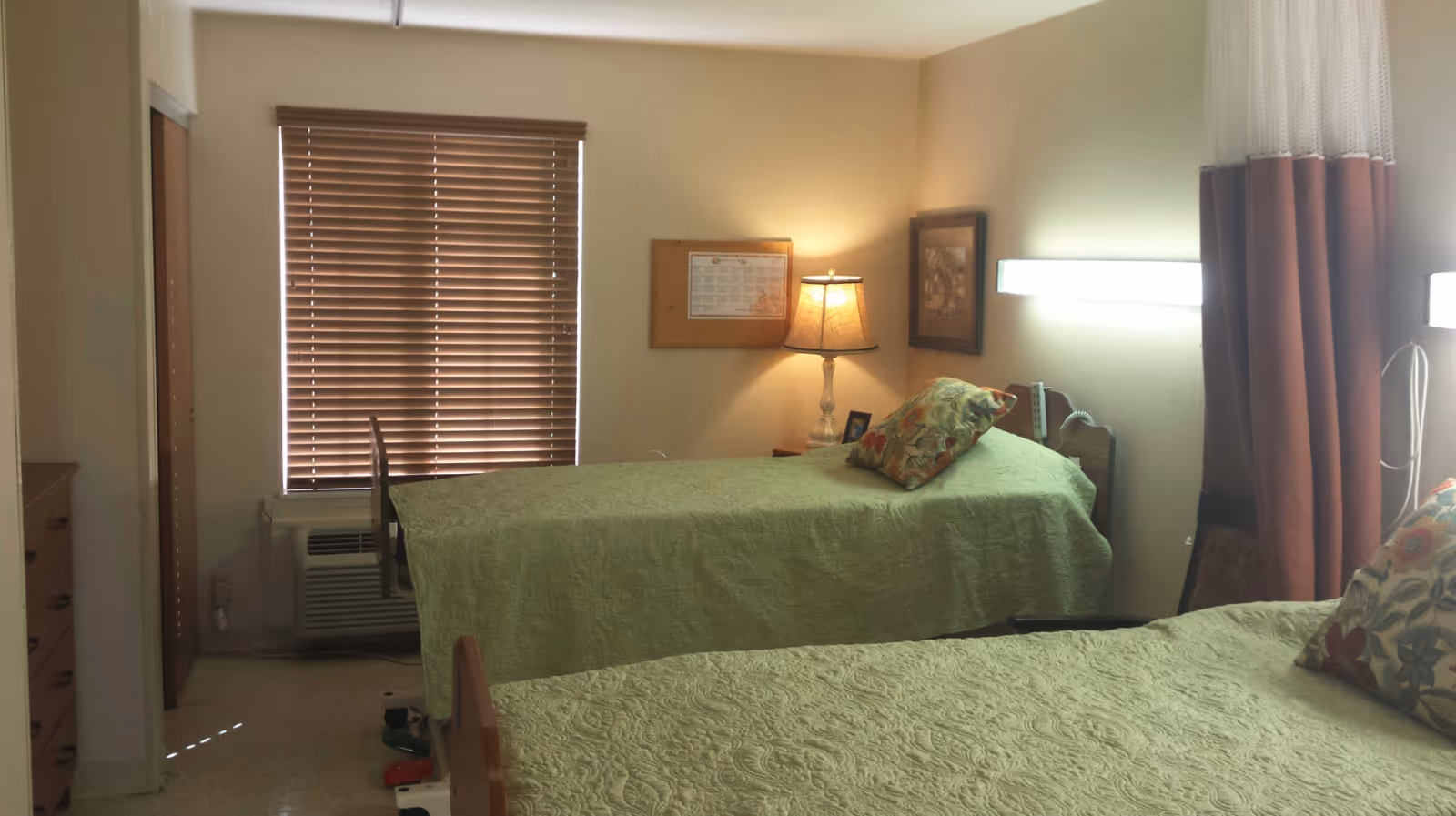A small, cozy bedroom with two single beds covered in light green bedspreads, each with a floral pillow. There is a window with closed wooden blinds, a lamp on a nightstand between the beds, a framed picture on the wall, and a curtain partially visible on the right side.