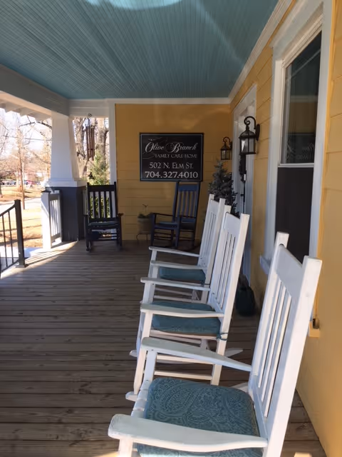 Front porch of a yellow house with white rocking chairs, a blue ceiling, and a sign for Olive Branch Family Care Home.