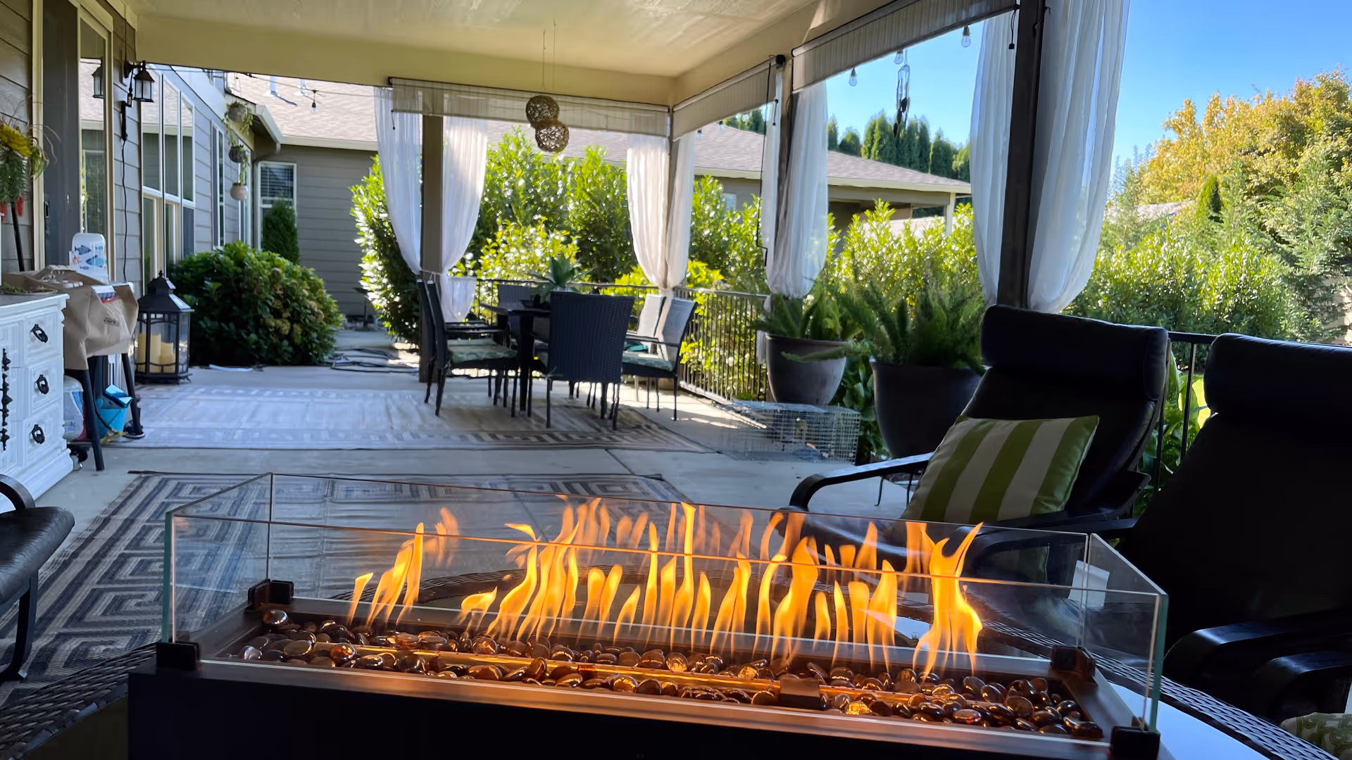 Covered outdoor patio with a glass-enclosed fire pit in the foreground, seating and a dining table under curtains and potted plants.