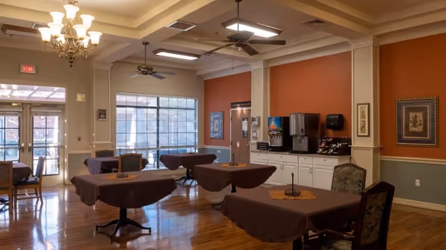 Interior view of a dining room in Keller Oaks Healthcare Center with several tables covered in brown tablecloths, chairs, a beverage station with coffee machines and cups, large windows letting in natural light, ceiling fans, and chandeliers.