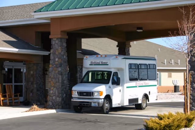 A white Highland Manor shuttle bus parked under the covered entrance of a senior living facility with stone columns and a green metal roof.