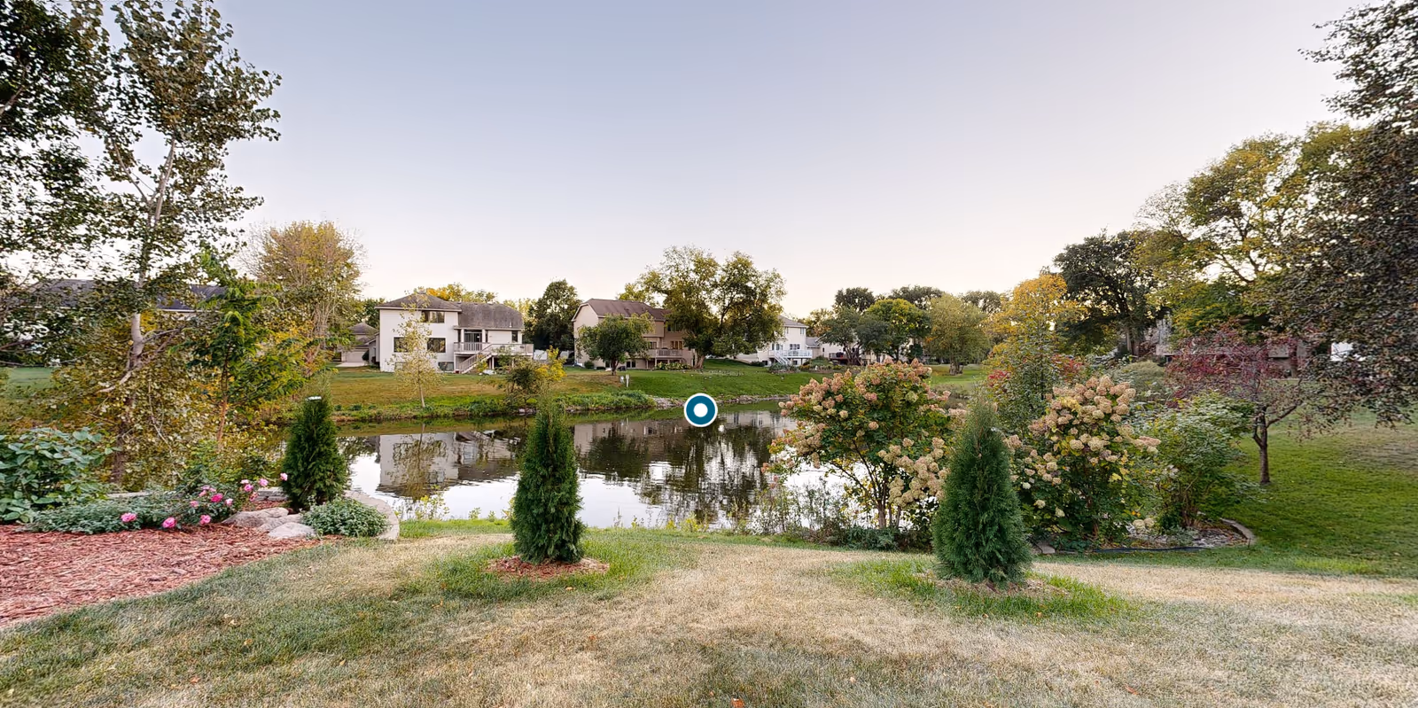 A peaceful outdoor scene featuring a small pond surrounded by green grass, bushes, and trees with houses visible in the background under a clear sky.