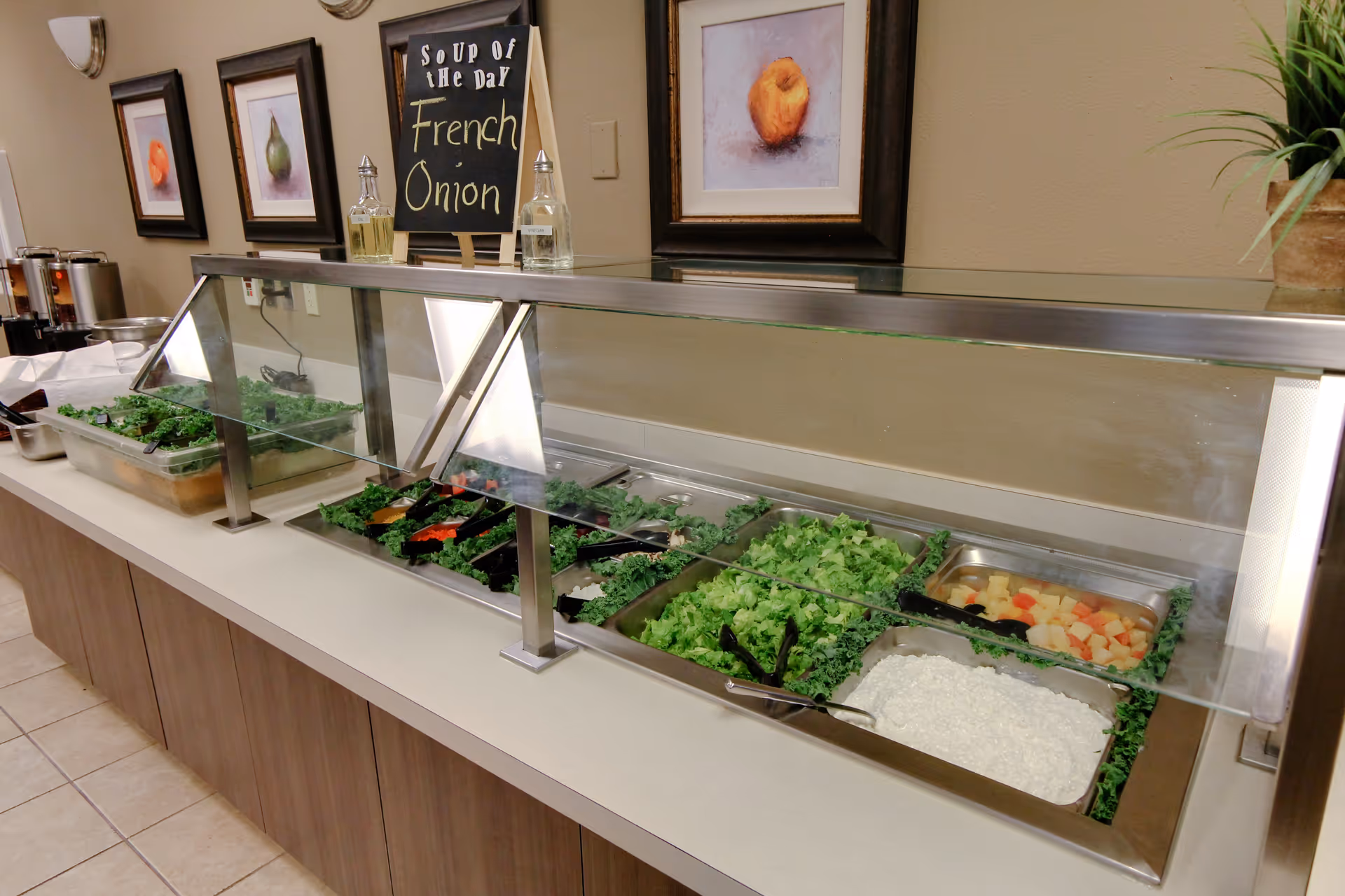 A salad bar with various fresh greens and toppings under a glass sneeze guard in a dining area. Behind the salad bar, there is a chalkboard sign that reads 'Soup of the Day French Onion' and framed pictures on the wall.
