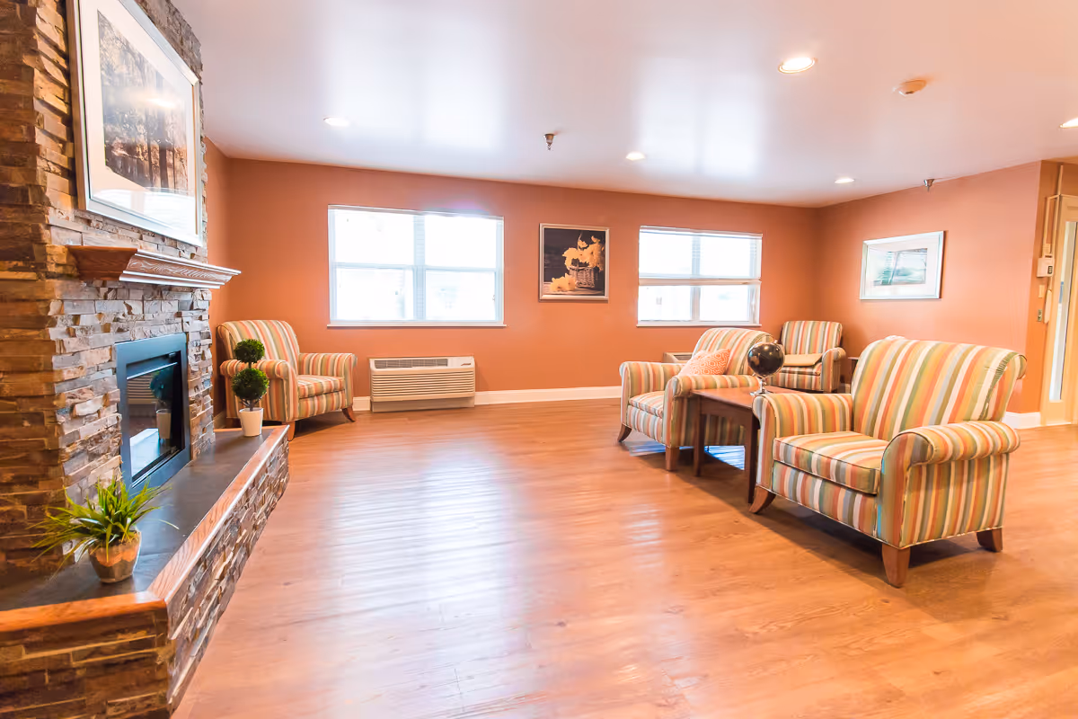 Bright communal living room with striped armchairs, a stone fireplace, hardwood floors and windows.