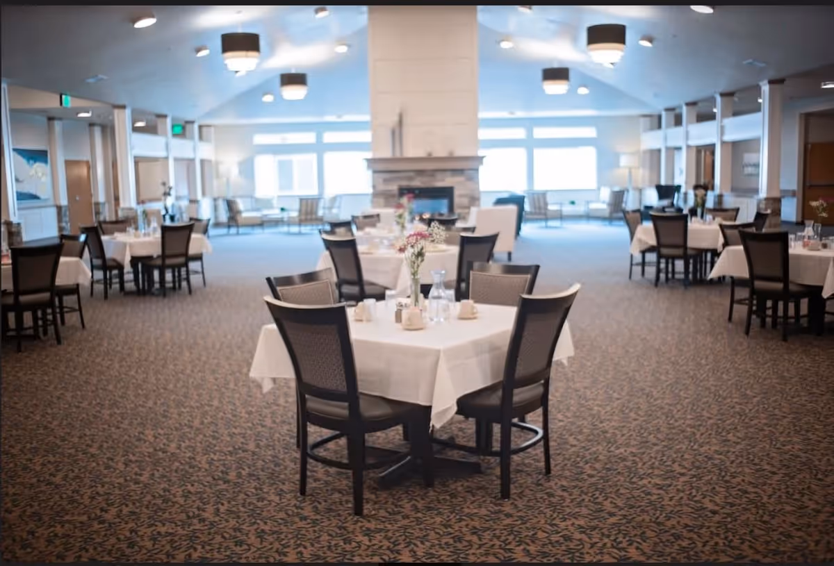 A spacious dining room with multiple tables covered in white tablecloths, each set with cups, glasses, and small flower arrangements. The room has carpeted floors, large windows letting in natural light, and a fireplace in the background. Comfortable chairs surround the tables, and there are seating areas with armchairs near the windows.