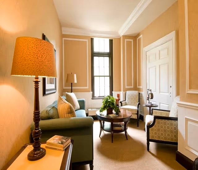 A cozy living room with beige walls and white trim, featuring a green sofa with striped pillows, two patterned armchairs, a round wooden coffee table with a plant and books, a floor lamp, and a window letting in natural light.