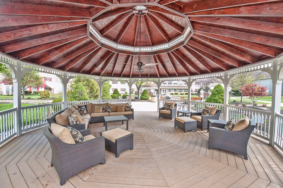 Spacious outdoor gazebo with a wooden ceiling and ceiling fan, furnished with cushioned wicker chairs, sofas, and ottomans arranged around a central area, surrounded by a white railing and overlooking a landscaped garden with trees and shrubs.