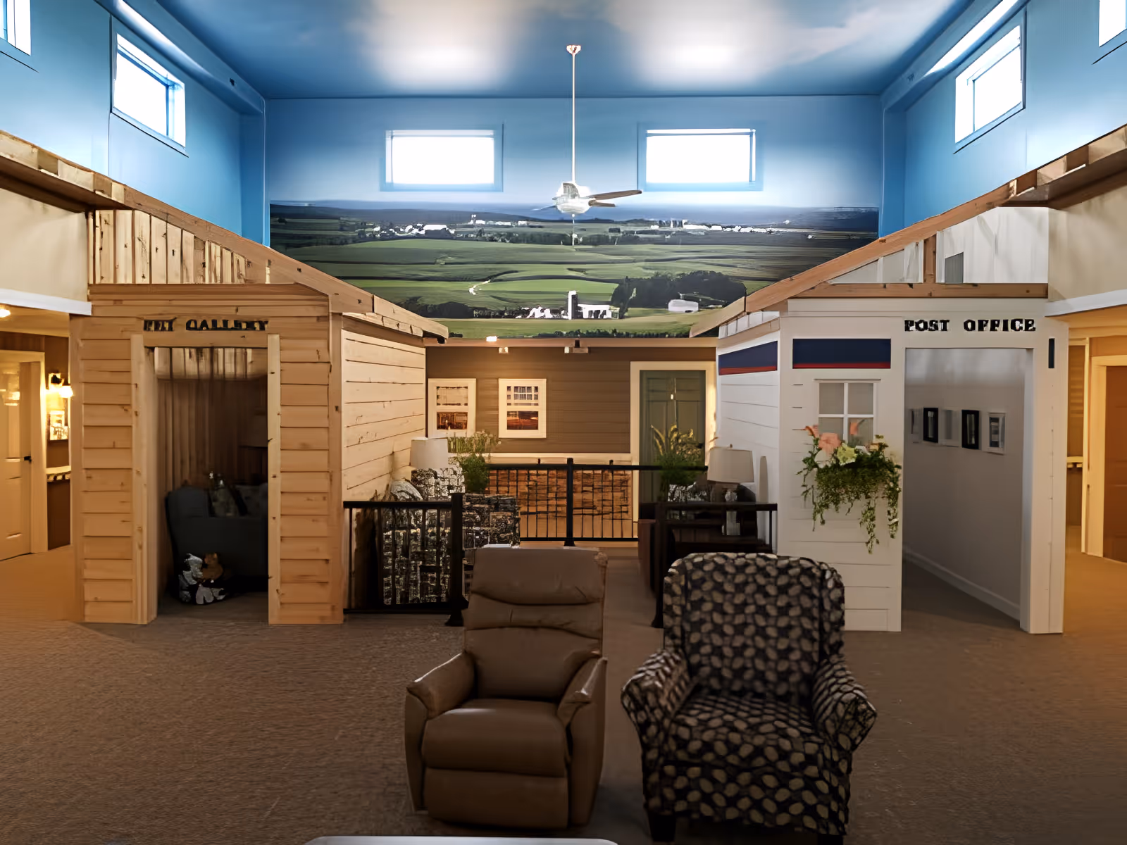 Indoor common area with two recliners in front of small mock storefronts labeled "Post Office" and "Art Gallery" beneath a blue ceiling mural.
