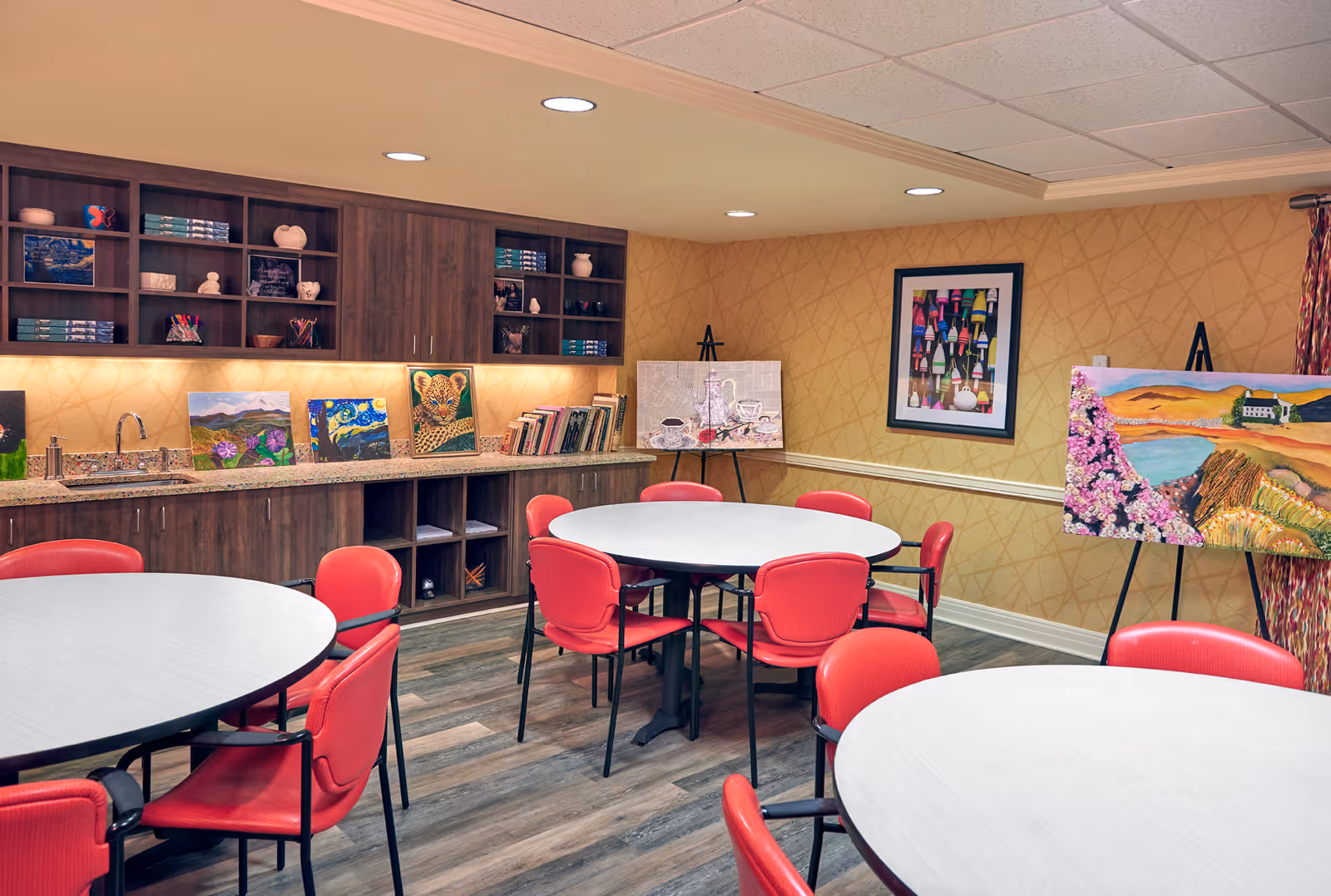 A well-lit room with round tables surrounded by red chairs. The back wall features wooden cabinets with shelves holding books, art supplies, and decorative items. Several colorful paintings are displayed on the countertop and on easels against the wall. The room has a warm, inviting atmosphere with patterned wallpaper and wood flooring.