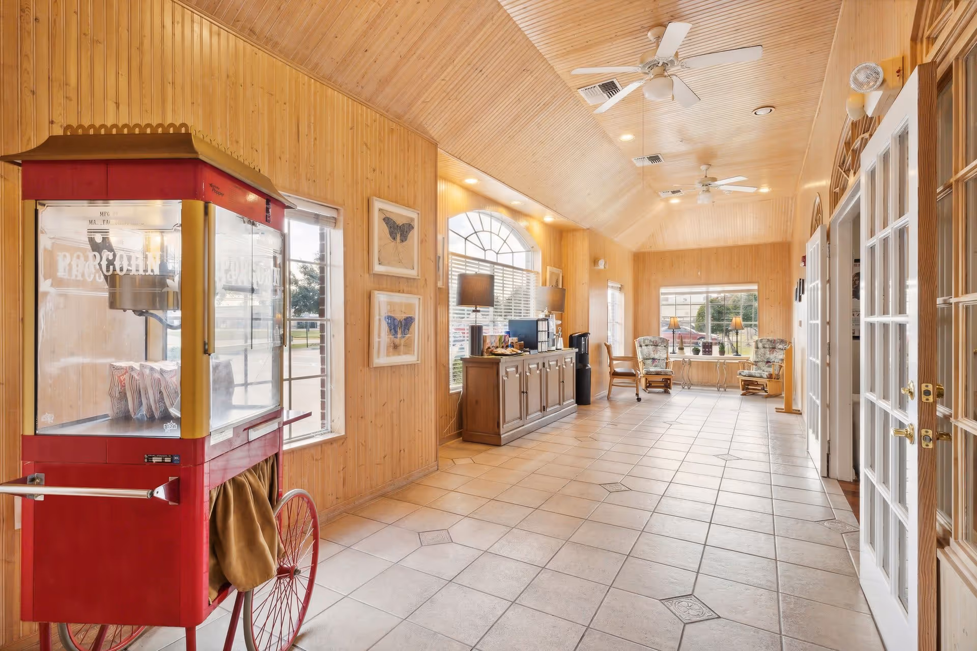 A bright hallway with wooden paneled walls and ceiling, tiled floor, and large windows letting in natural light. On the left side, there is a vintage red popcorn machine. Further down the hallway, there is a wooden cabinet with two lamps and a coffee station on top. At the end of the hallway, two cushioned chairs with floral upholstery are positioned near a window with two table lamps on a small table between them. Ceiling fans and recessed lighting are visible on the ceiling.