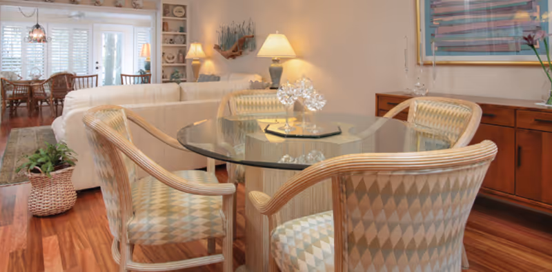 A cozy living room area with a round glass-top table surrounded by four cushioned chairs with a diamond pattern. Behind the table is a white sectional sofa, a wooden sideboard with decorative items, a lamp, and a framed artwork on the wall. In the background, there is a dining area with a wooden table and chairs near large windows with white shutters, allowing natural light to fill the space. The room has warm wooden flooring and a potted plant in a woven basket on the floor.