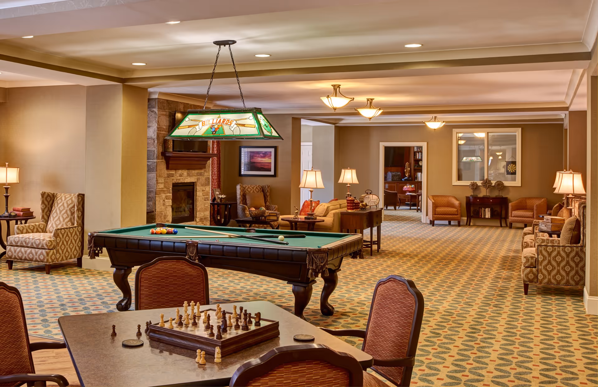 A spacious senior living lounge with a pool table, chess table, upholstered seating, lamps, and a fireplace.
