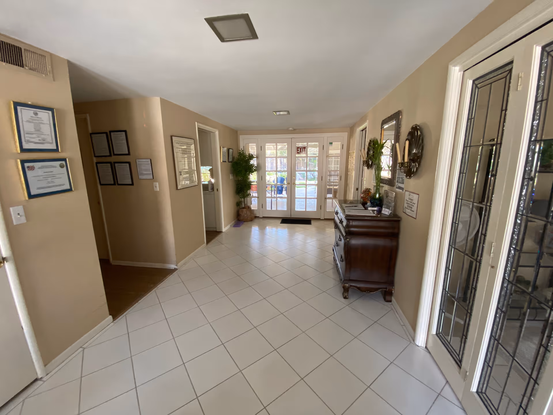 Bright tiled entry foyer with double glass doors, a wooden chest, framed certificates on the wall, and decorative plants.