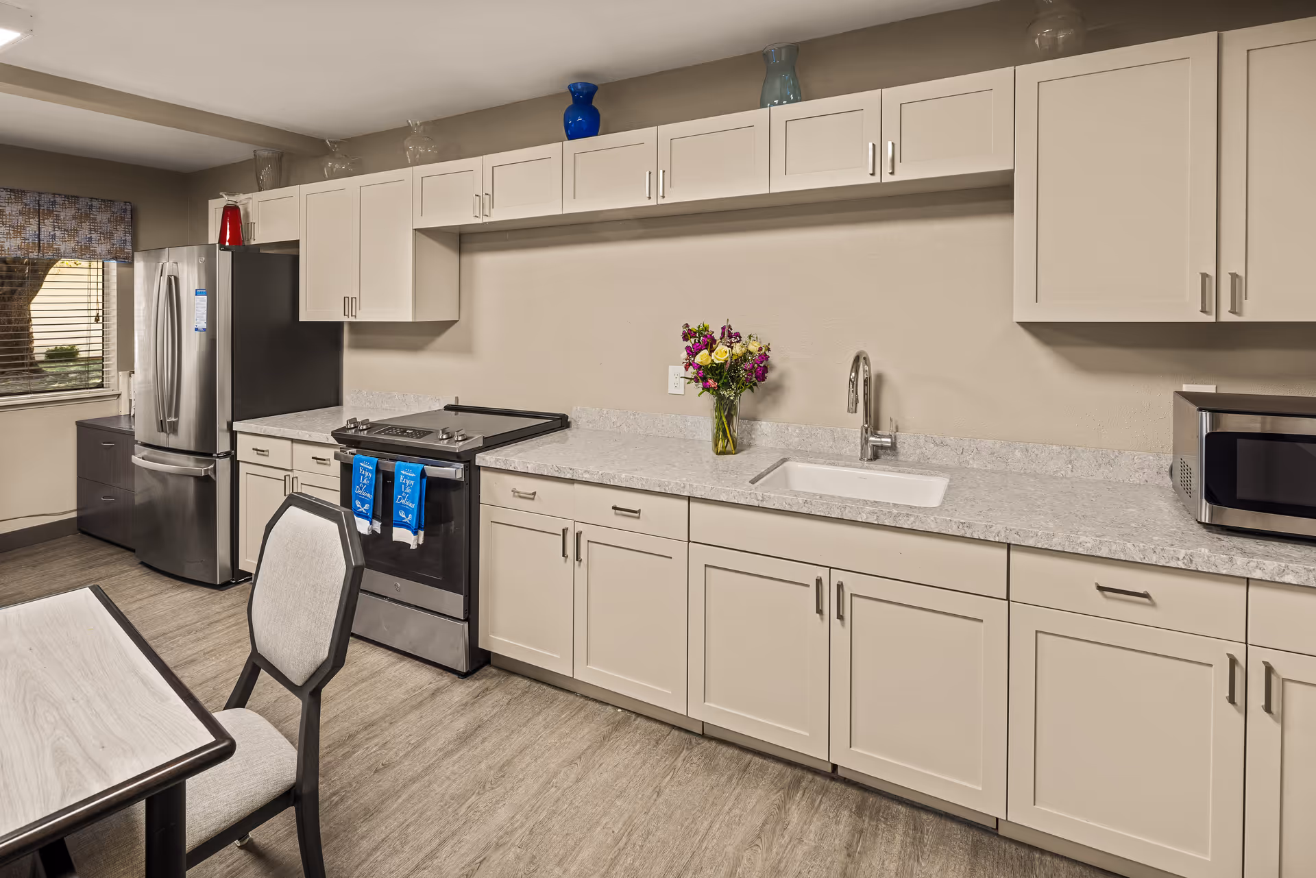 Modern kitchen area in a senior living facility featuring white cabinets, a stainless steel refrigerator, a stove with blue towels hanging on the handle, a sink with a faucet, a microwave on the countertop, and a vase with colorful flowers. There is a window with blinds and a table with a chair in the foreground.