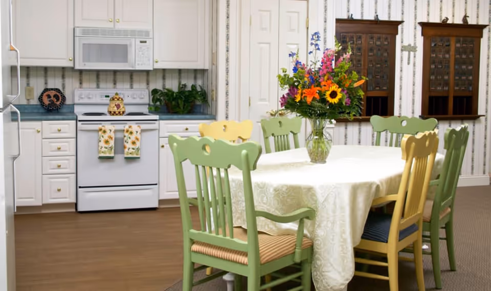 A dining area with a rectangular table covered with a white tablecloth and surrounded by six chairs painted in green and yellow. A vase with a colorful bouquet of flowers is placed in the center of the table. In the background, there is a kitchen area with white cabinets, a white stove with two floral towels hanging on the oven door, a microwave above the stove, and some plants on the countertop. The walls have a striped wallpaper with a floral pattern and two wooden mail organizers mounted on the wall.