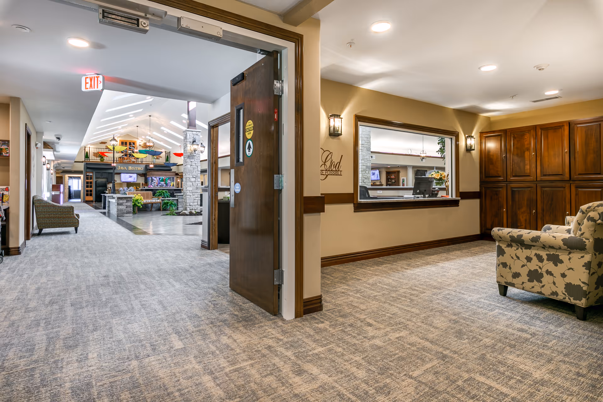 Interior view of a senior living facility hallway with carpeted floors, beige walls, and wooden trim. On the right side, there is a floral-patterned armchair and a window opening to a reception area with a computer. The hallway leads to a common area with a stone fireplace, plants, and a sign that reads 'Java Bistro'. The ceiling has skylights and recessed lighting, and there is an exit sign above a door on the left.