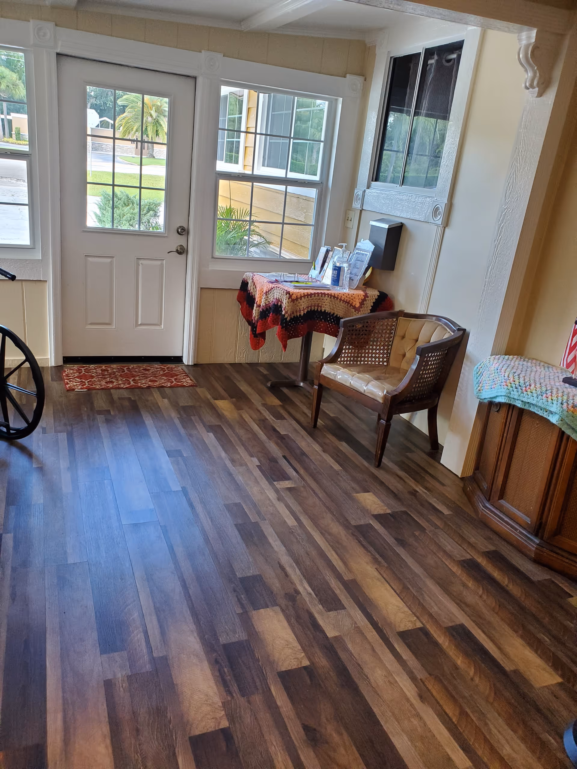 Interior view of a room with wooden flooring, a white door with glass panels, two windows, a small table covered with a colorful crocheted blanket holding various items, a wooden chair with a cushioned seat, and a wooden cabinet also covered with a crocheted blanket.