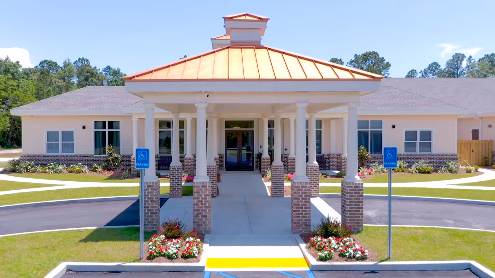 Front exterior view of The Homestead Assisted Living Community building with a covered entrance supported by white columns on brick bases, a copper-colored roof, landscaped flower beds, and two handicap parking signs near the driveway.