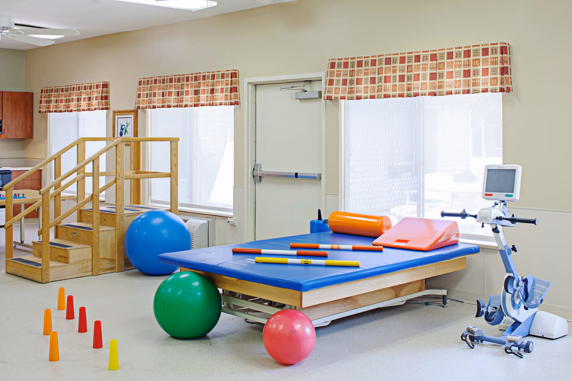 A physical therapy room with a blue padded therapy table holding various exercise equipment including foam rollers and rods. There are colorful exercise balls on the floor, a set of small orange, red, and yellow cones arranged in a line, a wooden staircase for mobility exercises, and a stationary exercise bike with a monitor. The room has large windows with patterned valances and light-colored walls.