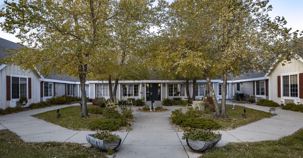 Courtyard entrance of a single-story assisted living facility with trees, paved walkways, planters, and outdoor seating.