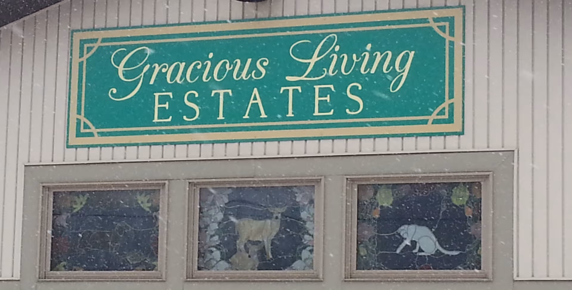 Exterior sign reading "Gracious Living ESTATES" mounted on a building above three decorative stained-glass windows.