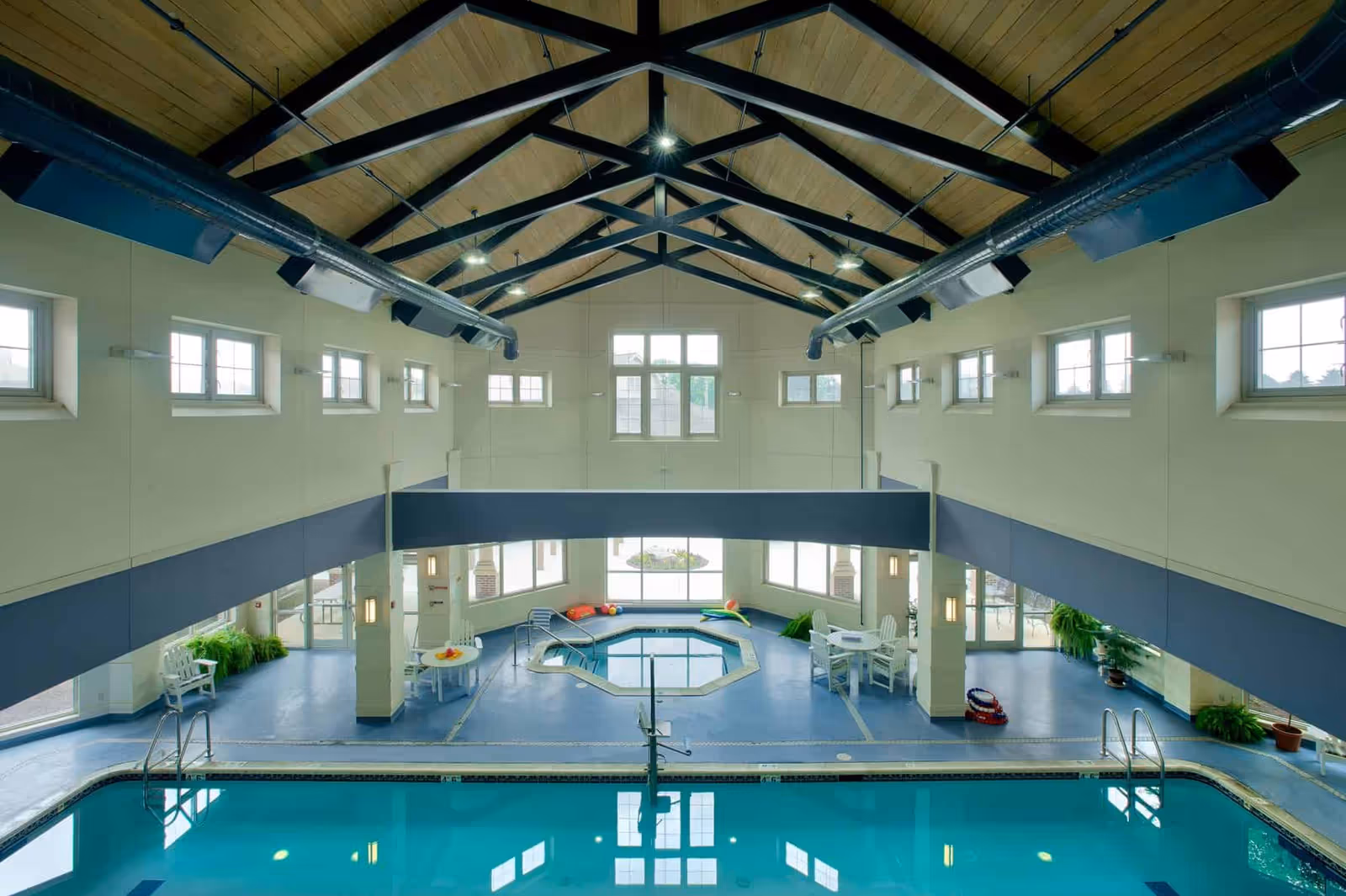 Indoor swimming pool area with a smaller hot tub in the background, surrounded by white chairs and tables. The room has high ceilings with exposed black beams and wooden panels, multiple windows letting in natural light, and potted plants placed around the perimeter.
