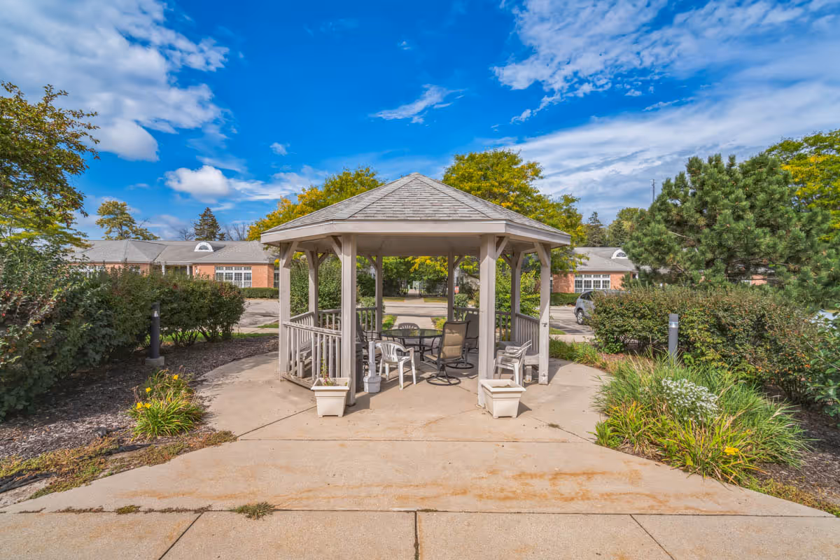 Outdoor gazebo with a hexagonal roof surrounded by chairs and a table, set on a concrete patio with greenery and bushes around. In the background, there are buildings and a parking area under a partly cloudy blue sky.