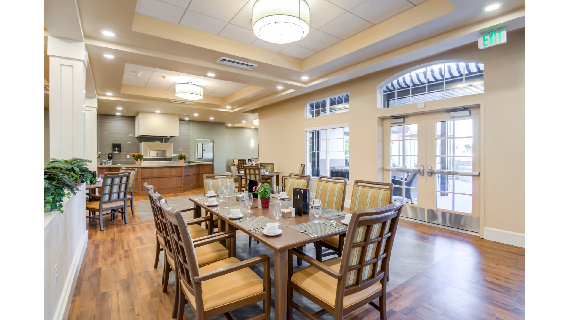 A bright and spacious dining room in a senior living facility with wooden floors, a large dining table set with placemats, cups, glasses, and a small potted plant centerpiece. The room features multiple chairs with cushioned seats and backs, large windows and glass doors leading outside, and a kitchen area with modern appliances and cabinetry in the background.