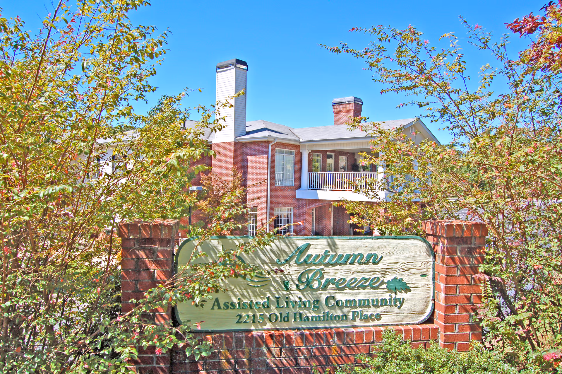 Front exterior of a brick assisted living building with trees and a wooden sign reading 'Autumn Breeze Assisted Living Community'.