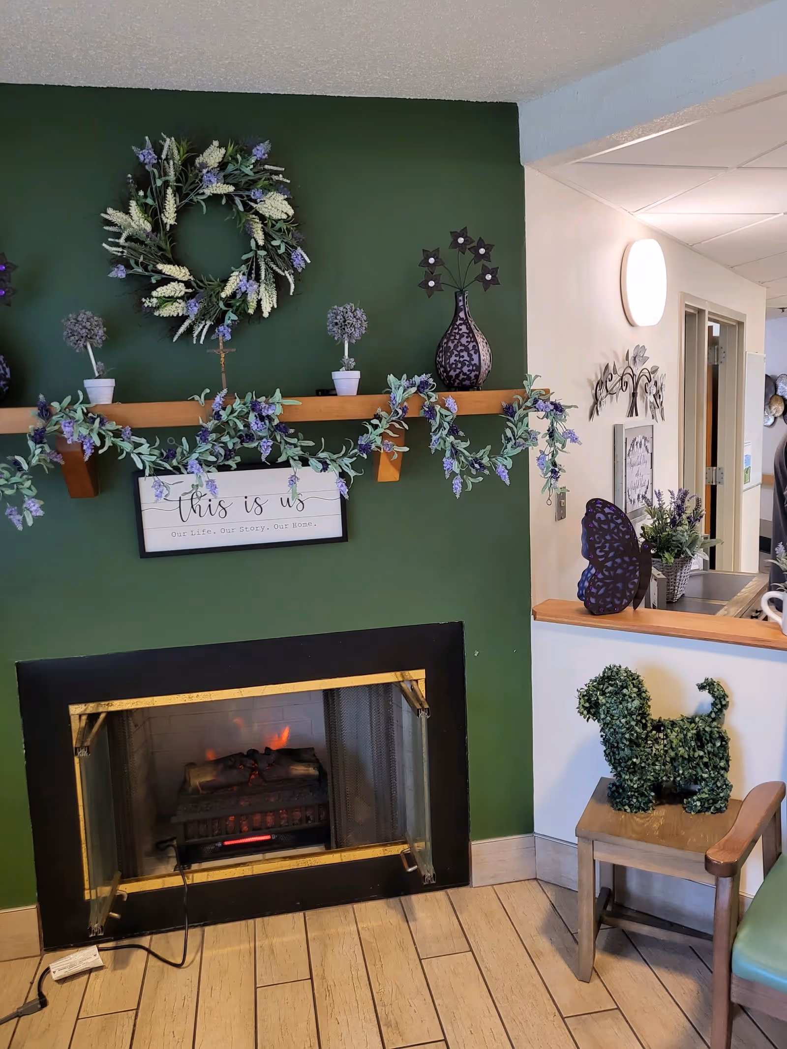 Cozy interior corner with a green accent wall featuring a fireplace below a wooden shelf decorated with a floral wreath, small potted plants, and a sign that reads 'this is us Our Life, Our Story, Our Home'. To the right, a small wooden chair holds a decorative topiary shaped like a dog, and a counter with a purple butterfly decoration and a flower arrangement is visible.