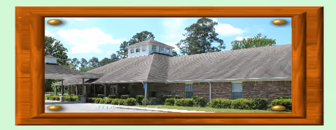 Exterior view of a single-story brick building with a sloped roof and a small tower-like structure on top, surrounded by greenery and trees under a partly cloudy sky. The image is framed with a wooden border.