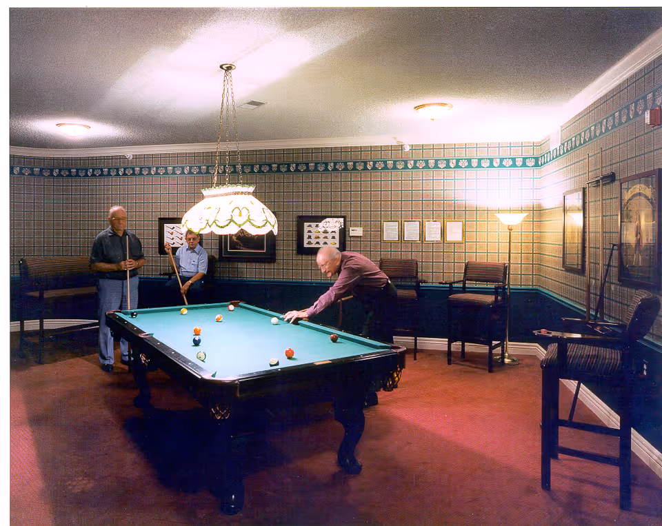 Three elderly men playing pool in a room with patterned wallpaper and red carpet. The room is furnished with a pool table, chairs, framed pictures on the walls, and a hanging light fixture above the pool table.
