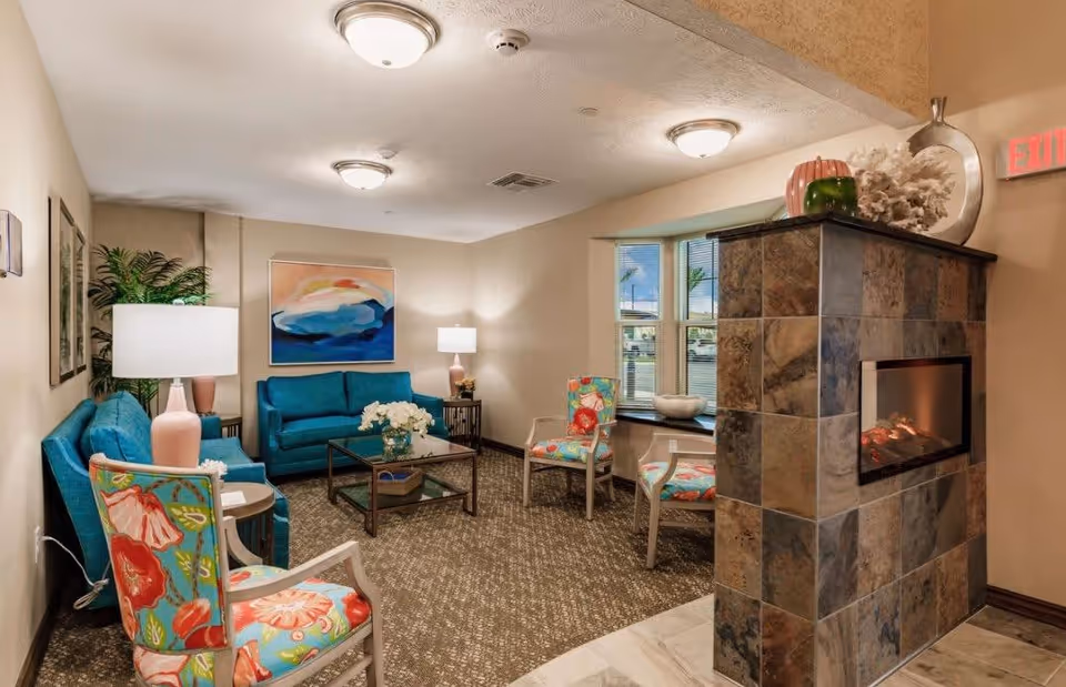 A cozy living room area in a senior living facility featuring two teal sofas, three floral-patterned chairs, a glass coffee table with a flower arrangement, two table lamps, a large abstract painting on the wall, a tiled fireplace with decorative items on top, and a window with a view outside.