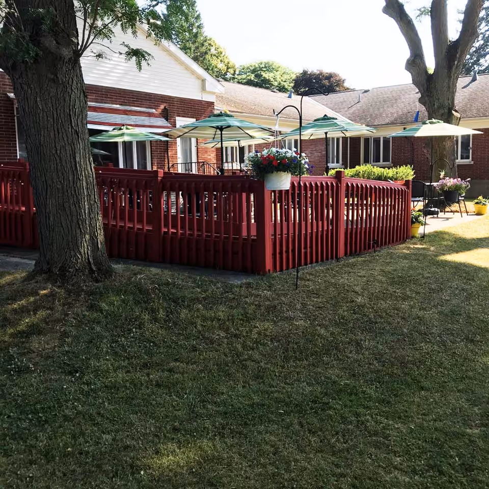 Outdoor patio area at Westminster Manor with red wooden fencing, several green and white striped umbrellas providing shade over tables and chairs, hanging flower baskets, and large trees surrounding the space.
