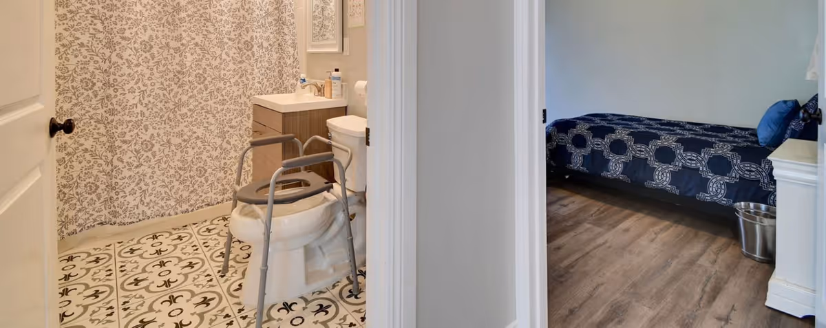 View of a bathroom with patterned tile floor, a shower with a floral curtain, a toilet with a raised seat and support frame, and a sink with a mirror above it. Adjacent to the bathroom is a bedroom with a single bed covered in a dark blue patterned bedspread and a small metal trash can on the wooden floor.