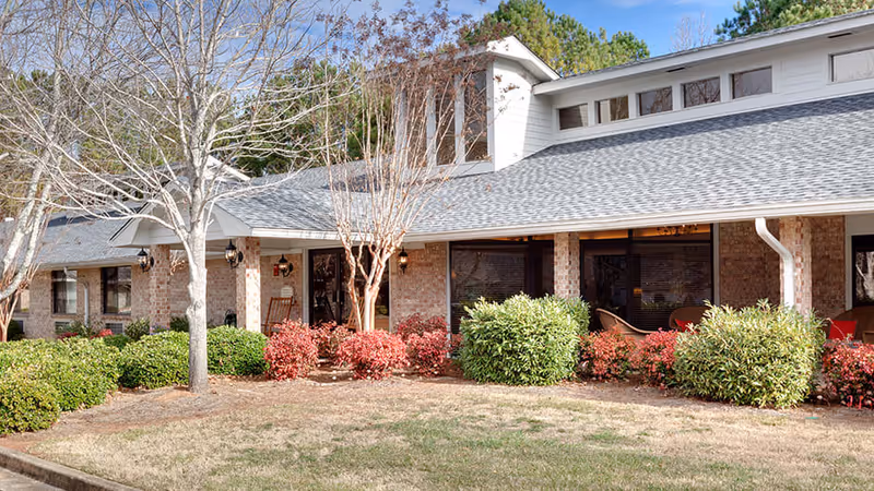 Exterior view of a single-story brick building with a gray shingled roof, surrounded by trimmed bushes and small trees with some red foliage, under a clear blue sky.
