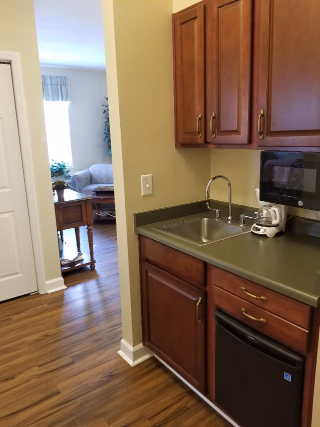Small kitchenette with wooden cabinets, sink, microwave and mini-fridge opening into a living room.