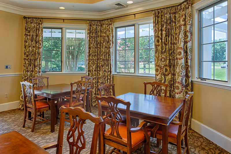 A dining room with wooden tables and chairs featuring orange cushions. The room has large windows with patterned curtains, allowing natural light to brighten the space. Outside the windows, greenery and trees are visible.
