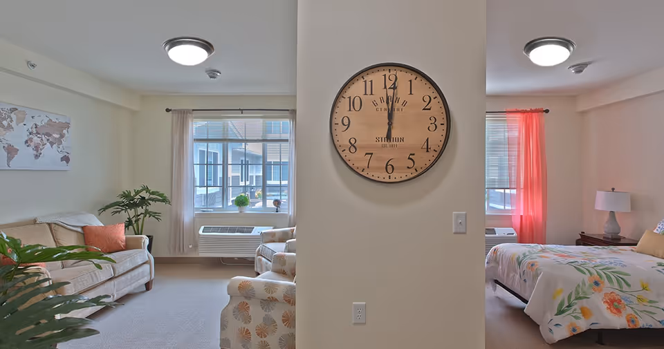 Interior view of a senior living facility room divided by a wall with a large round clock. On the left side is a living area with a beige sofa, two patterned armchairs, a potted plant, and a window with light curtains. On the right side is a bedroom area with a bed covered in floral bedding, a nightstand with a lamp, and a window with pink curtains.