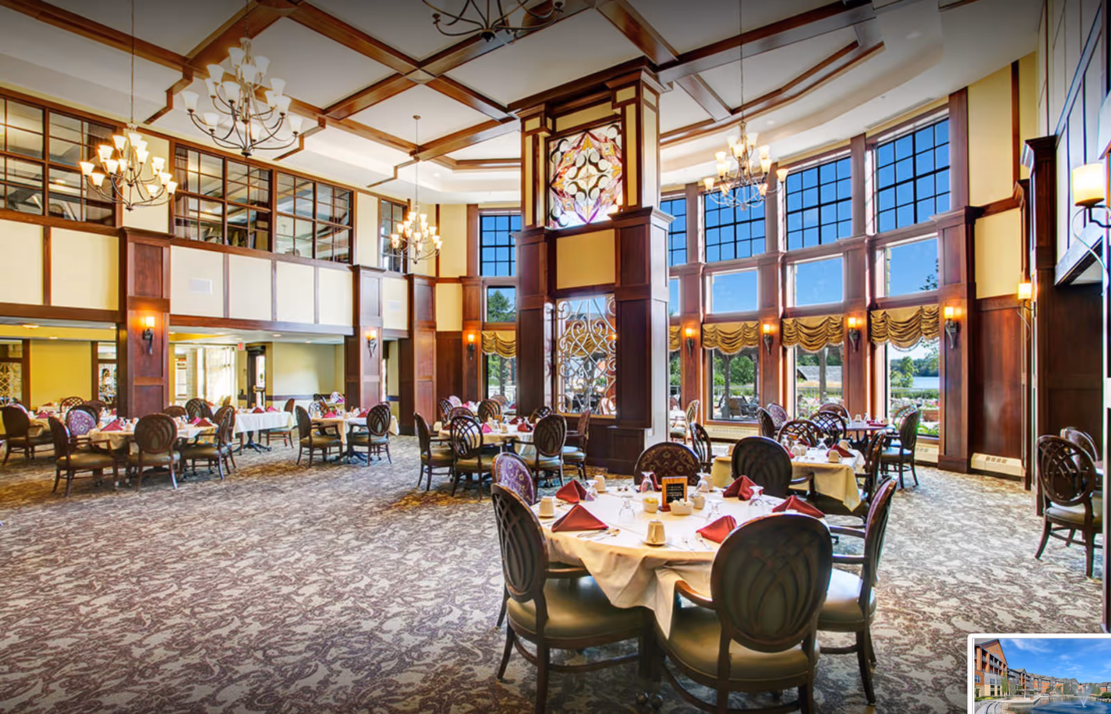 A spacious and elegant dining room with high ceilings, large windows letting in natural light, round tables set with white tablecloths, red napkins, and dining ware. The room features wooden paneling, chandeliers, and patterned carpet, with a decorative column in the center and views of greenery and water outside.