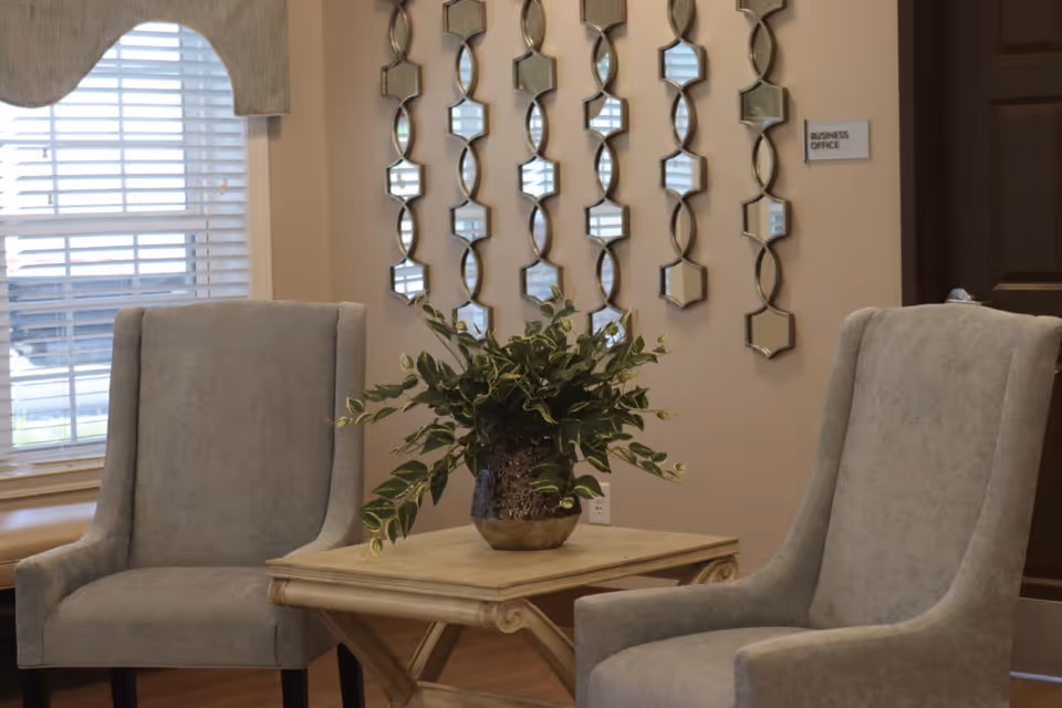 Two gray upholstered chairs flank a wooden side table with a potted plant in a decorated sitting area.